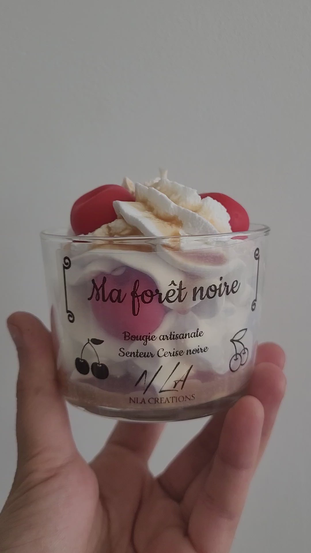 Vidéo gros plan montrant une bougie artisanale parfum Cerise Noire Intense, style forêt noire en pot, chantilly blanche avec cerise rouge brillante et coulis marron, révélant les détails gourmands et faits main.