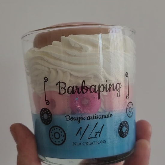 Vidéo en gros plan d’une bougie artisanale tenue dans la main, aux couleurs bleu pâle et jaune pâle, décorée de petits doigts rose fuchsia et surmontée d’un gros donuts rose vif, tournée doucement pour dévoiler tous ses détails gourmands et colorés.