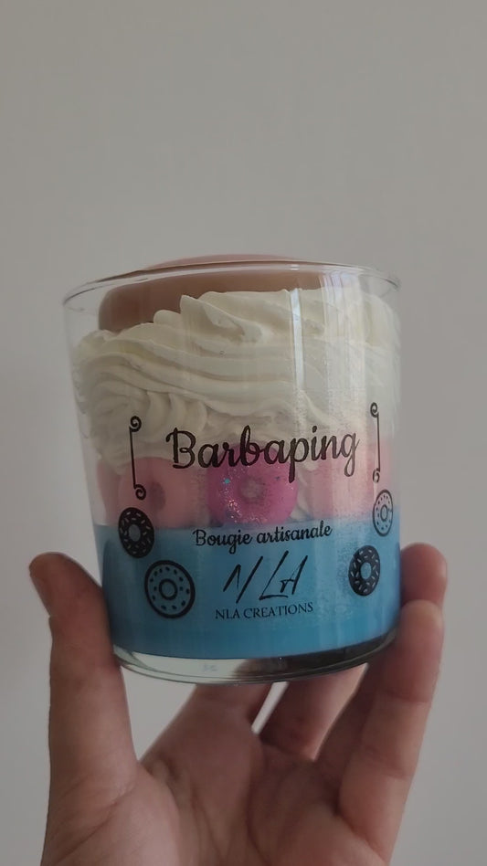 Vidéo en gros plan d’une bougie artisanale tenue dans la main, aux couleurs bleu pâle et jaune pâle, décorée de petits doigts rose fuchsia et surmontée d’un gros donuts rose vif, tournée doucement pour dévoiler tous ses détails gourmands et colorés.