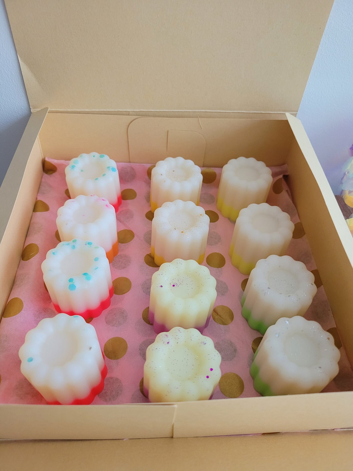 Coffret ouvert contenant 12 fondants parfumés en forme de petits cannelés, disposés sur un papier rose à pois dorés. Les fondants présentent différentes couleurs et décorations, avec des nuances de blanc, jaune, rose et des paillettes. Le coffret repose sur une surface en bois clair, à côté d'une rose décorative iridescente.