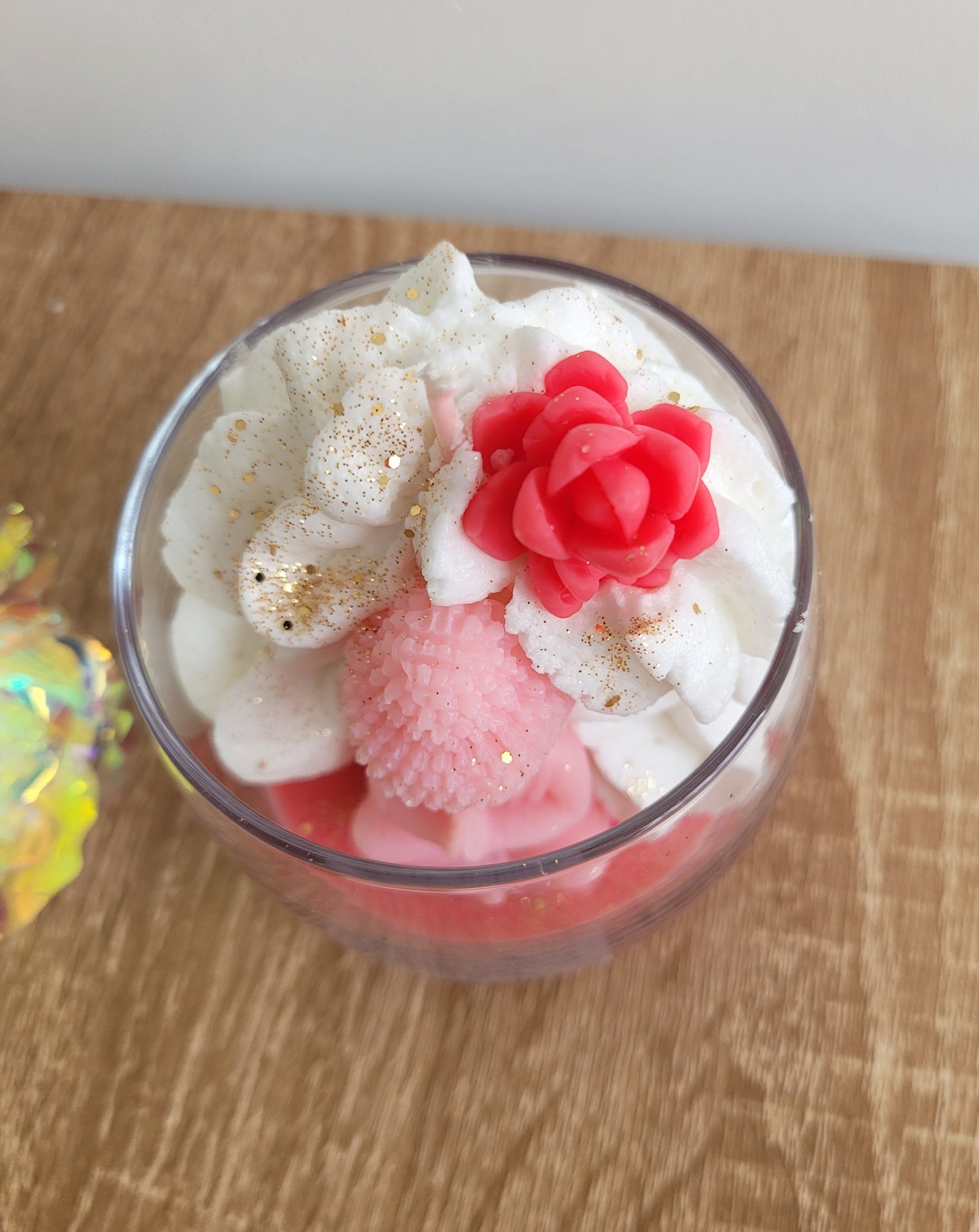 Vue plongeante sur une bougie rose à la fleur de cerisier, avec chantilly de cire fouettée, figurine Bouddha centrale et paillettes dorées scintillantes.