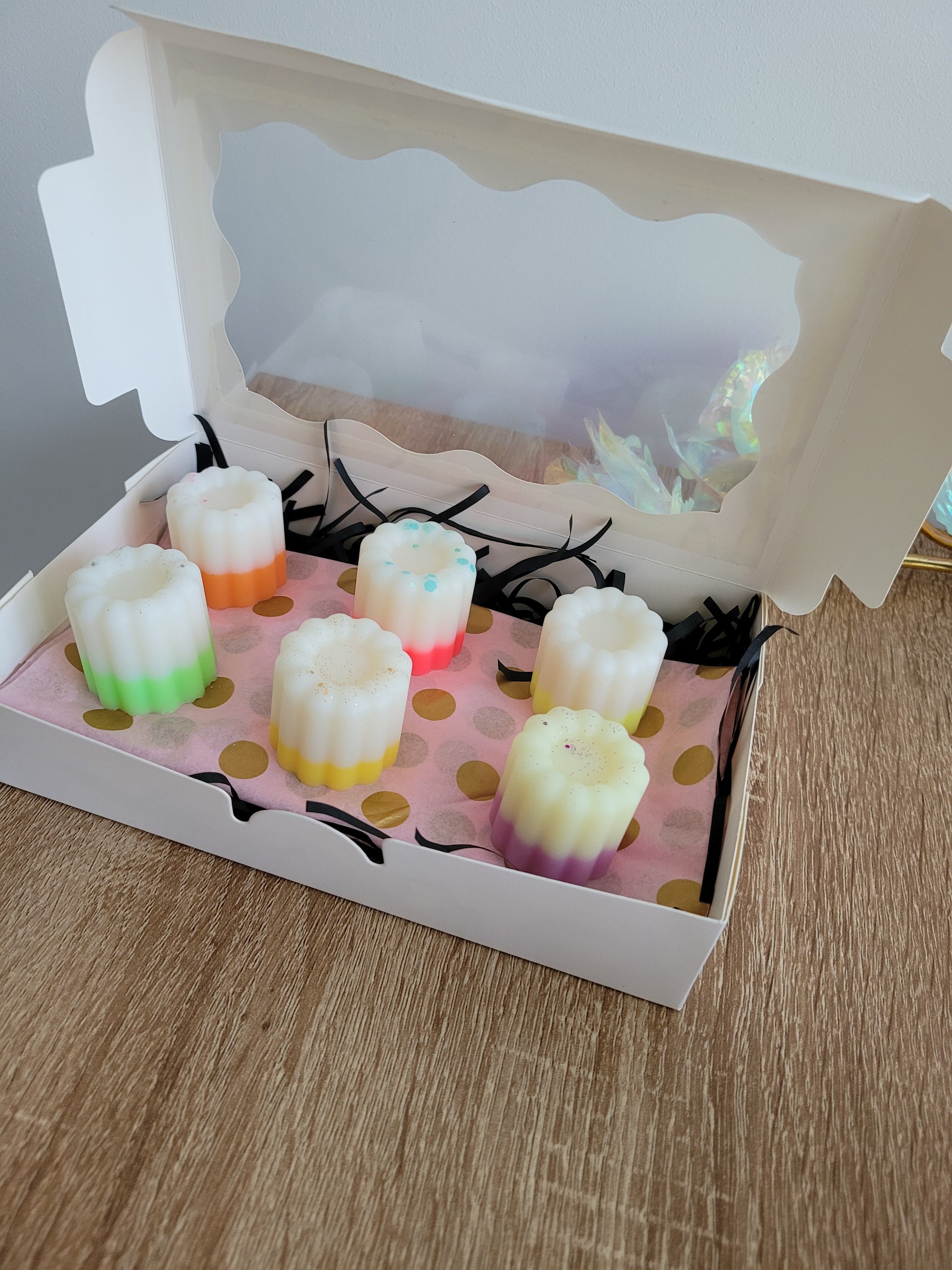 Boîte blanche ouverte contenant 6 fondants parfumés artisanaux en forme de cannelés, aux couleurs pastel et dégradés colorés (vert, rose, jaune, violet). Les fondants sont présentés sur un papier rose à pois dorés, avec des brins de raphia noir décoratif. Le couvercle transparent laisse entrevoir l’intérieur. La boîte repose sur une surface en bois clair, à côté d’une rose décorative iridescente.