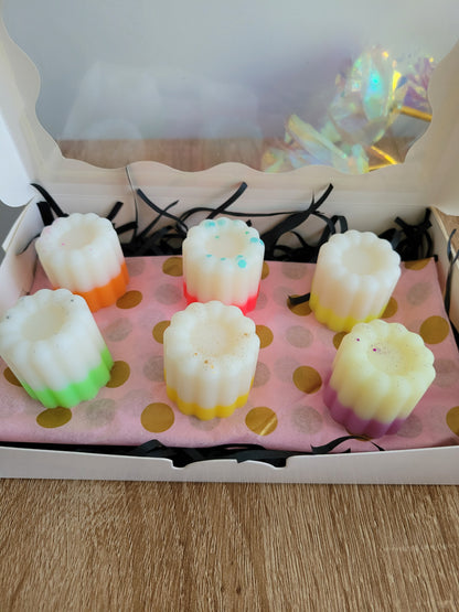 Boîte de 6 fondants parfumés en forme de cannelés, aux couleurs douces et dégradées, posés sur un papier rose à pois dorés. Chaque fondant a une teinte différente, et la boîte blanche est décorée de brins noirs. L’ensemble repose sur une table en bois.