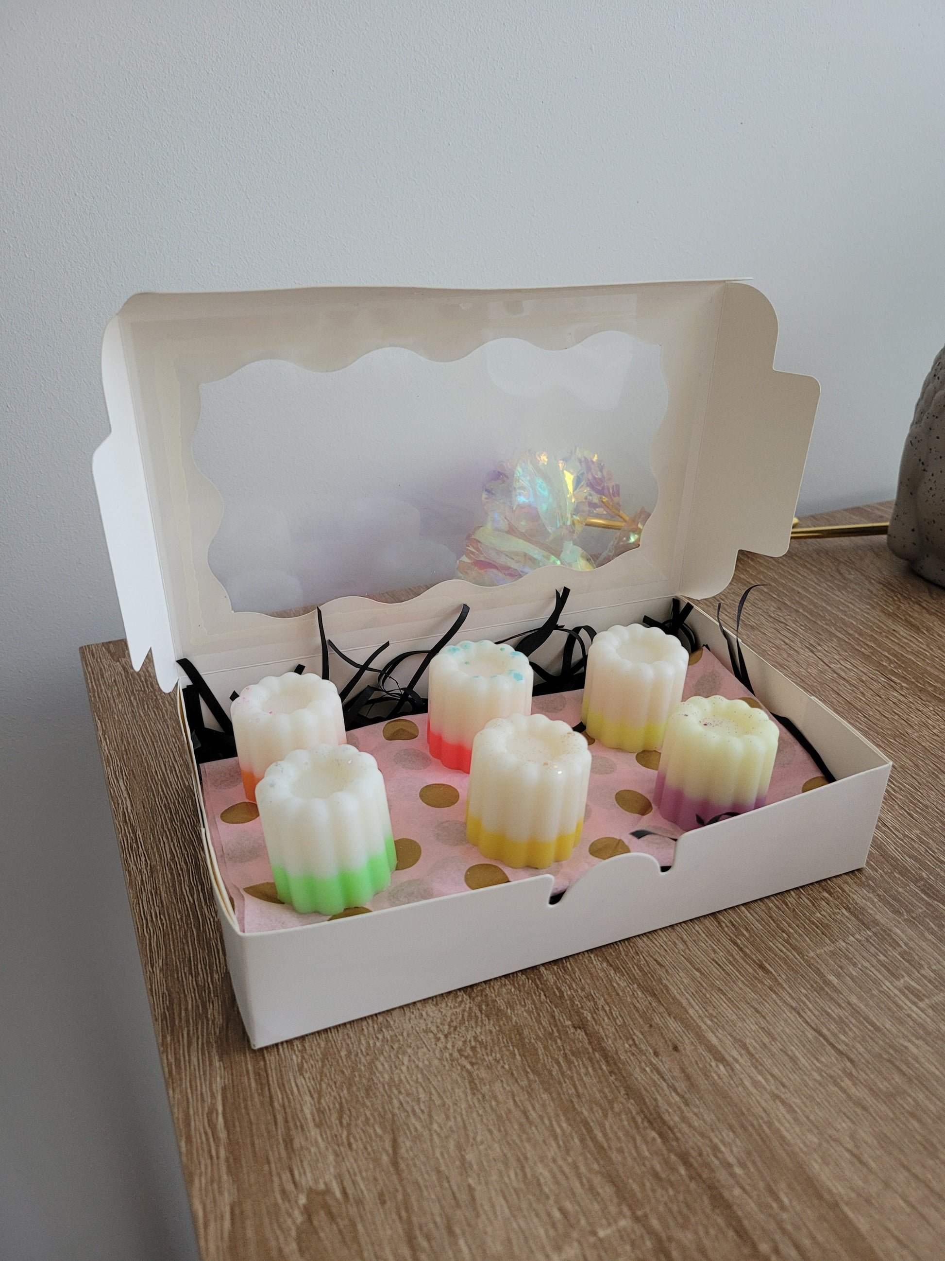 Fondants parfumés faits main en forme de cannelés, présentés dans une élégante boîte blanche avec fenêtre transparente. Le coffret contient 6 fondants aux couleurs estivales (rose, vert, jaune, violet), décorés de paillettes, sur un fond rose à pois dorés. Idéal pour offrir ou se faire plaisir avec des senteurs artisanales.