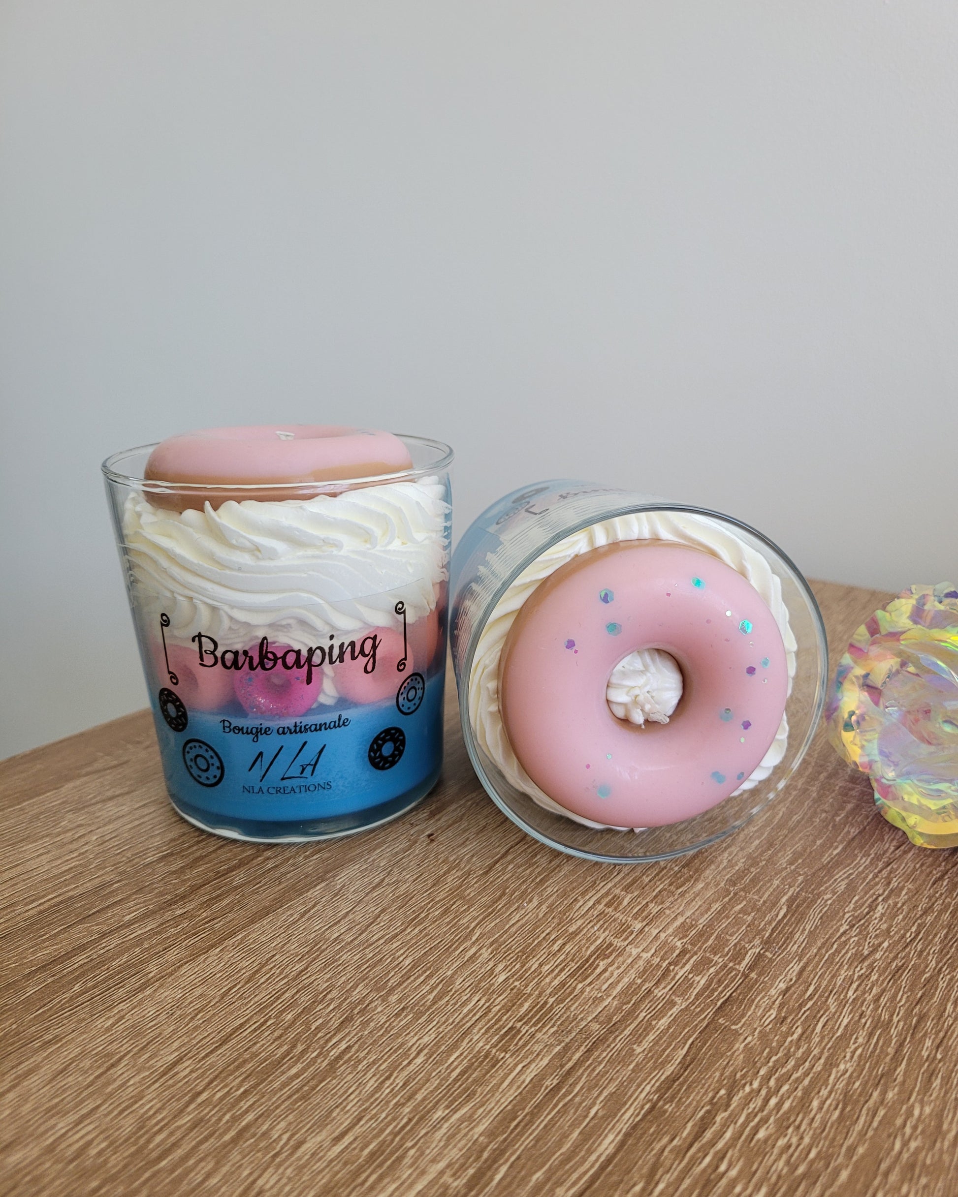 Photo barbe à papa prise du dessus d’une bougie artisanale aux teintes bleu pâle et jaune pâle, ornée de petits doigts rose fuchsia tout autour et surmontée d’un gros donuts rose vif au centre, mettant en valeur la texture et les détails colorés de la décoration.
