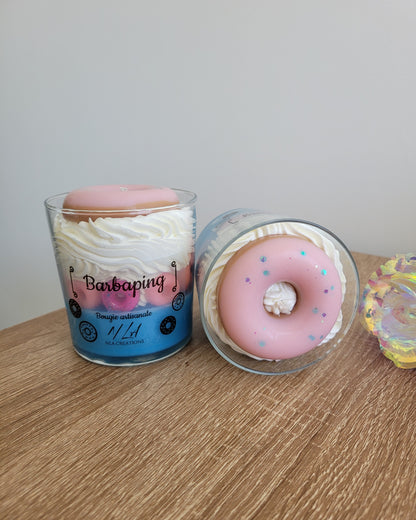 Photo barbe à papa prise du dessus d’une bougie artisanale aux teintes bleu pâle et jaune pâle, ornée de petits doigts rose fuchsia tout autour et surmontée d’un gros donuts rose vif au centre, mettant en valeur la texture et les détails colorés de la décoration.
