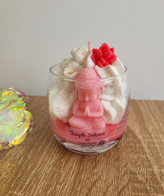 Gros plan frontal sur une bougie rose à la fleur de cerisier, décorée de chantilly de cire, d’une figurine Bouddha et de paillettes dorées brillantes.