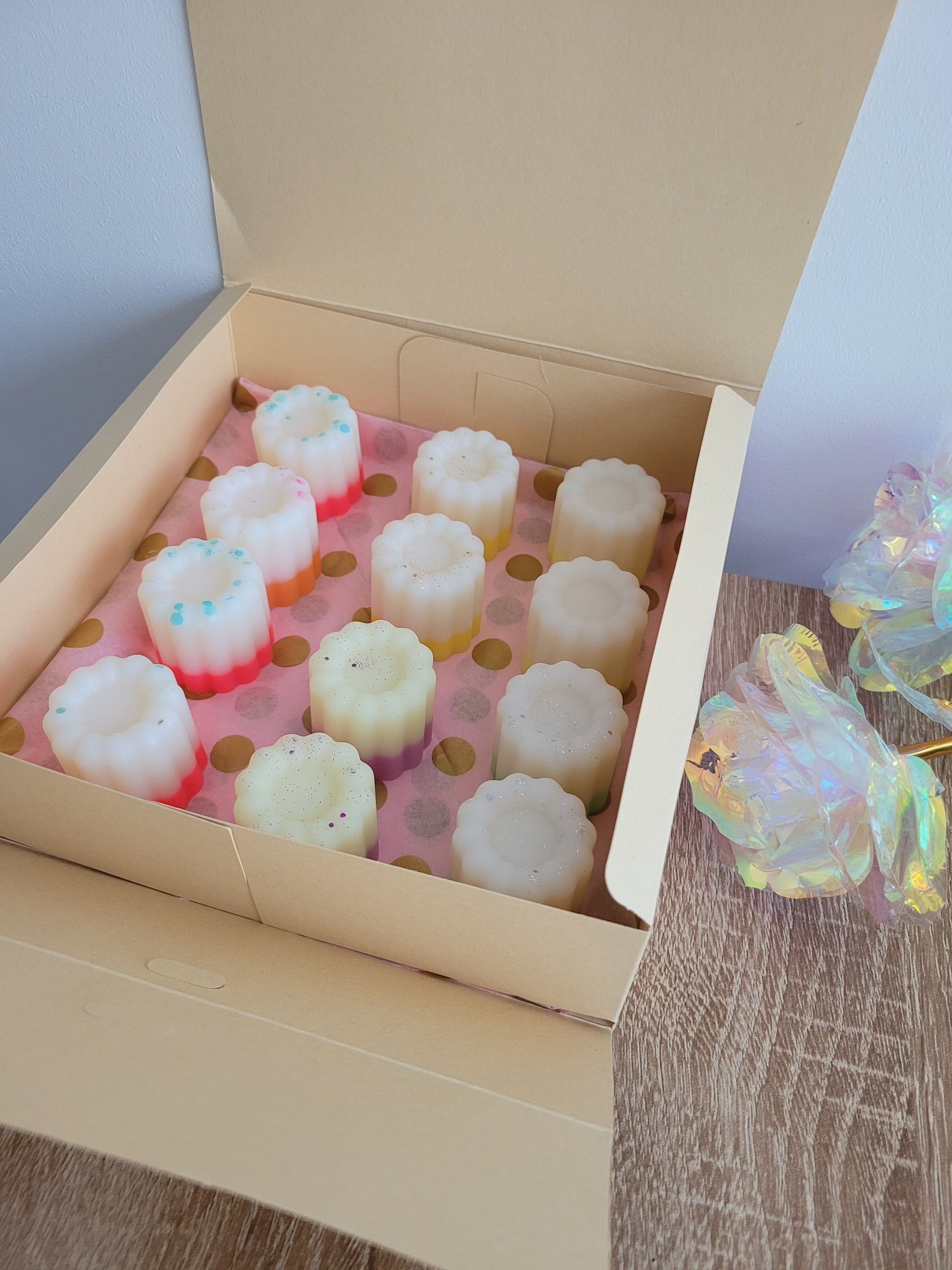 Boîte ouverte contenant 12 fondants parfumés artisanaux en forme de cannelés, posés sur un papier rose à pois dorés. Chaque fondant présente des couleurs douces et des décorations pailletées, avec des dégradés de blanc, rose et jaune. La boîte est posée sur une table en bois clair, à côté d'une rose décorative iridescente.